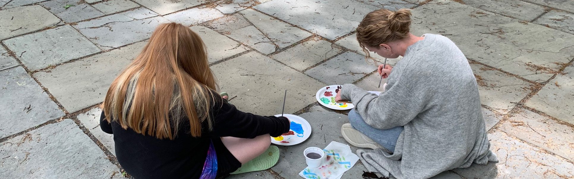 image of two students sitting outside on the ground and painting.